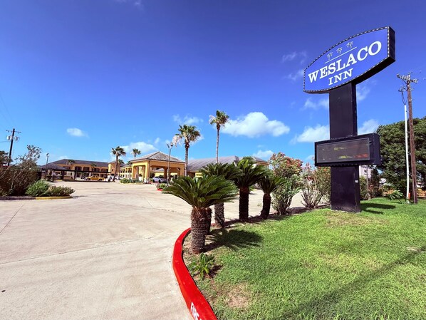 A hotel sign, a parking lot, and a building with a tiled roof. - Weslaco Inn Weslaco/Mercedes (Weslaco)