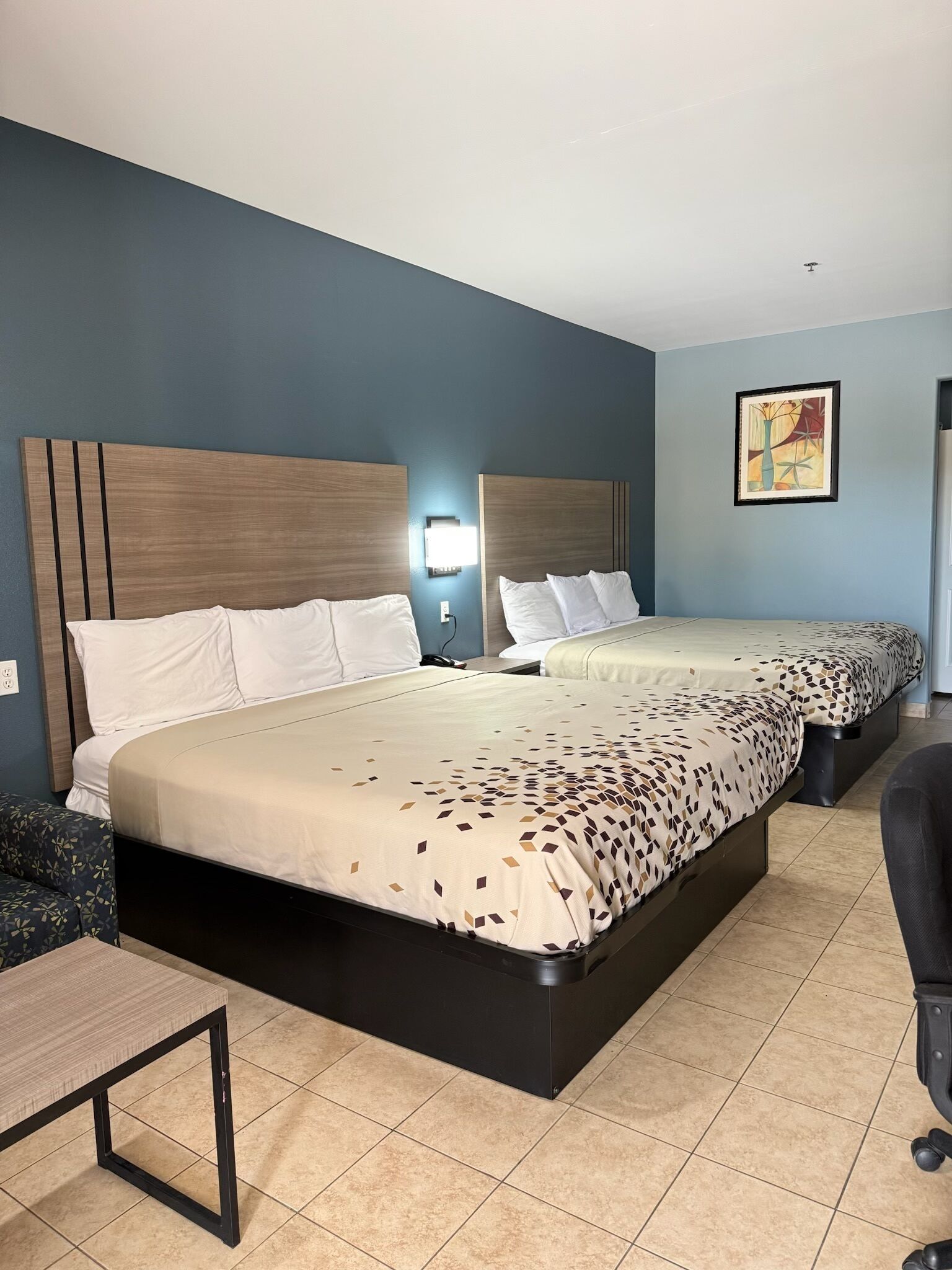 A hotel room with two beds, a desk, a chair, and a wall-mounted light.