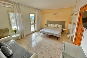 A hotel room with a bed, a sofa, a small table with a vase, a TV, and a balcony with curtains.