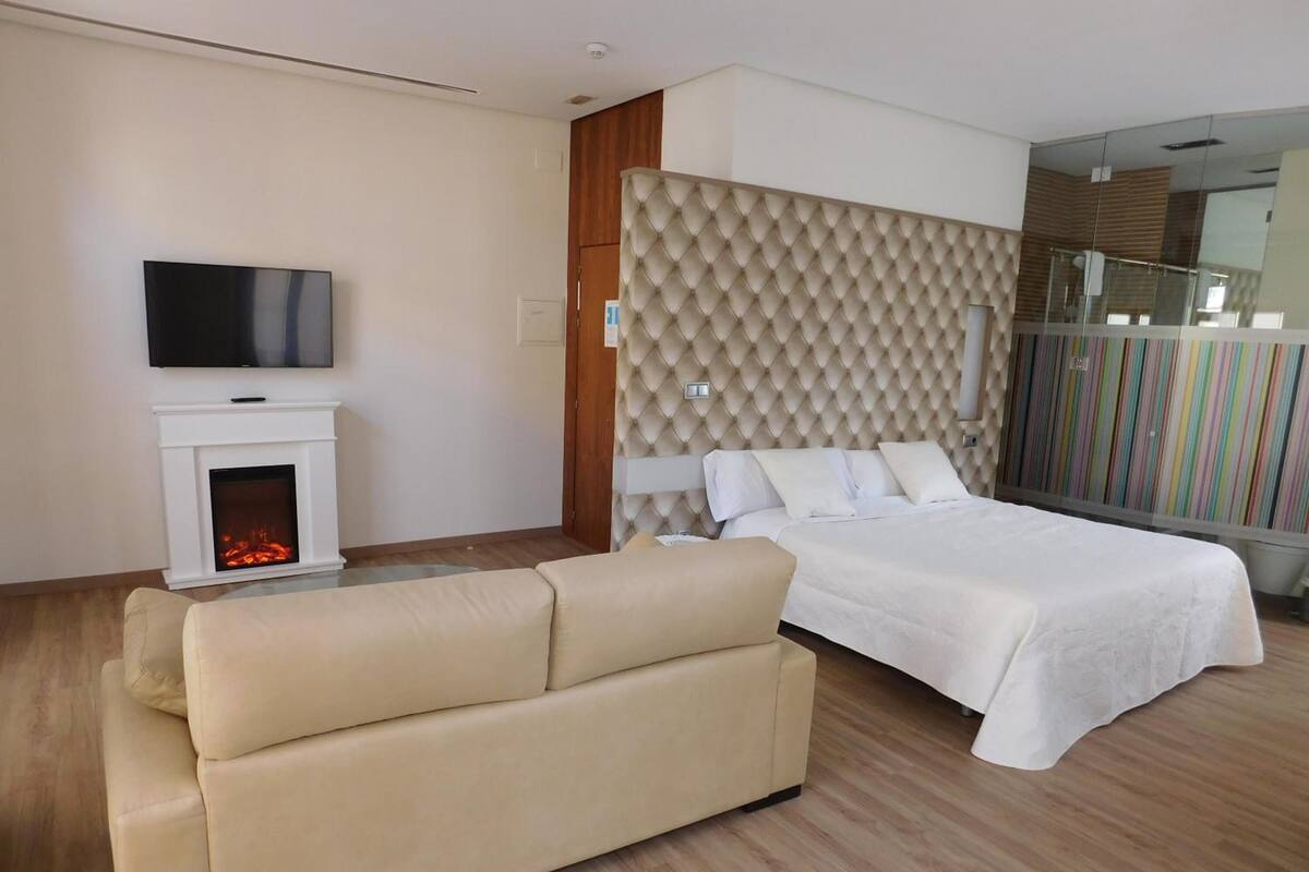 A modern hotel room with a large bed, a sofa, a fireplace, and a television.