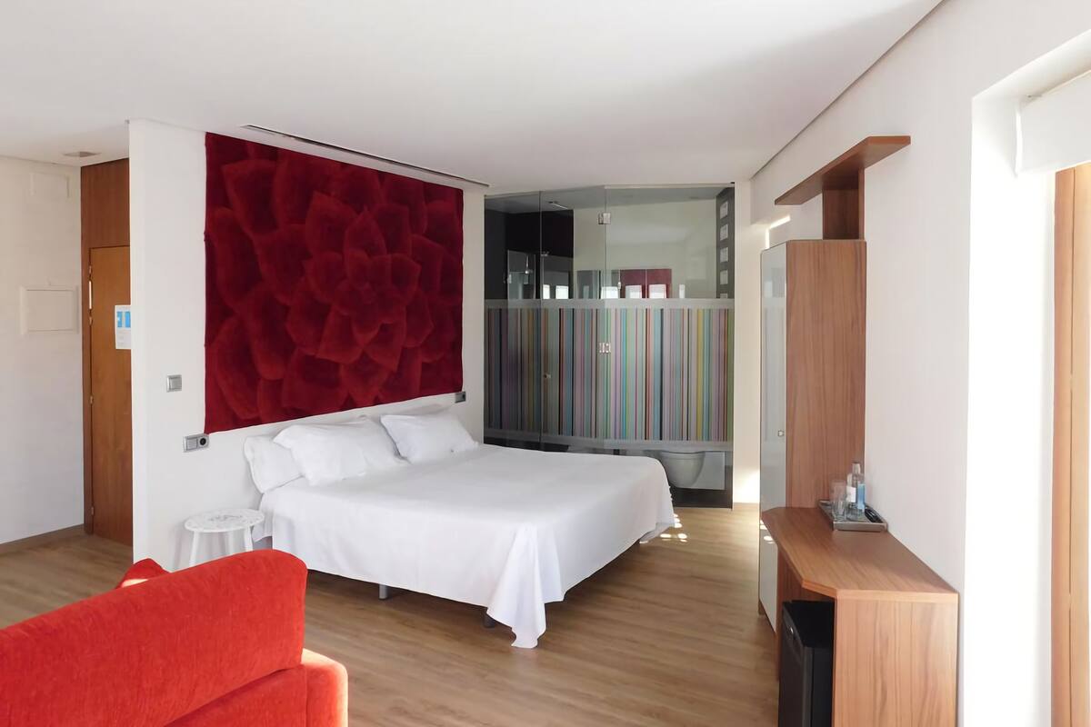 A modern hotel room with a large bed, a red accent wall, a small table, and a wardrobe with colorful stripes.