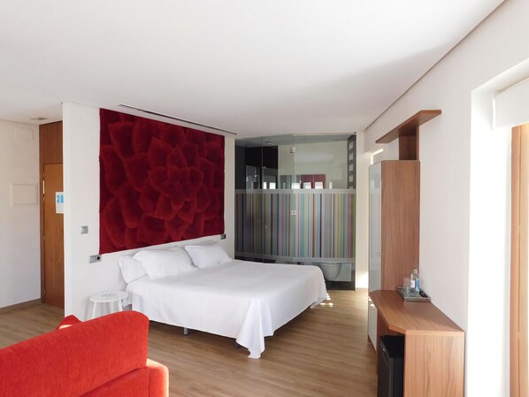 Moderni hotellihuone, jossa on suuri sänky, punainen tehosteseinä, pieni pöytä ja värikkäillä raidoilla koristeltu vaatekaappi.