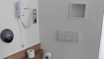 Un baño moderno con inodoro, secamanos automático, dispensador de toallas de papel, cubo de basura y un estante pequeño con una caja de pañuelos.