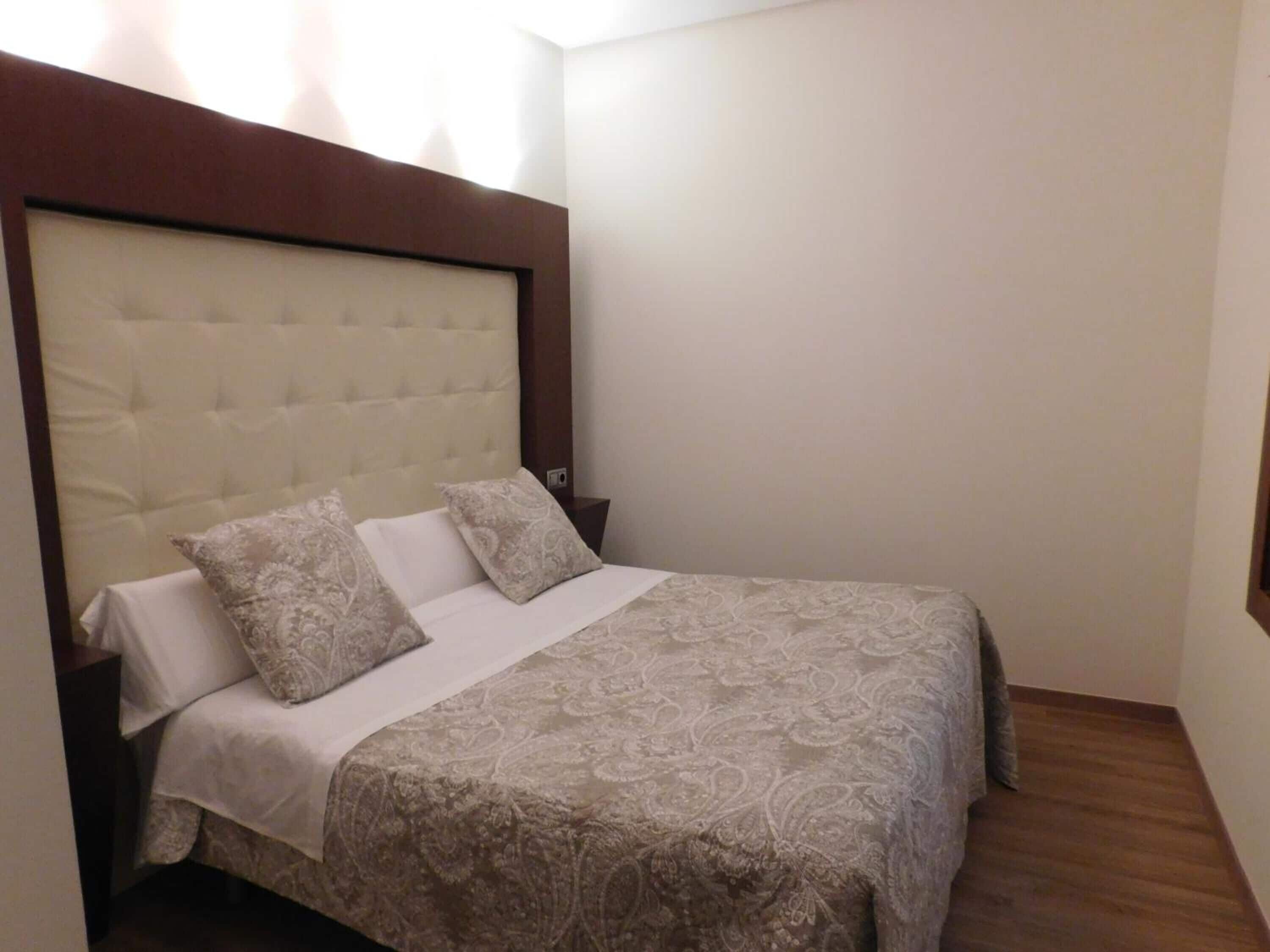 A neatly made bed with a tufted headboard, two pillows, and a patterned bedspread in a room with wooden flooring and plain walls.