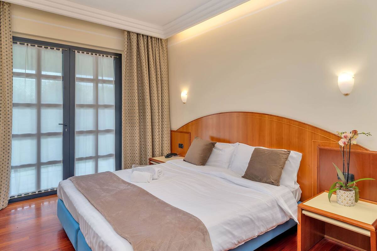 A hotel room with a bed, a bedside table with a lamp, a window with curtains, and a door.