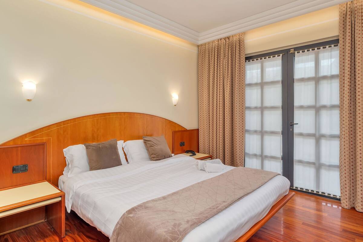 A hotel room with a wooden headboard, a bed with white and beige bedding, a bedside table, a phone, and a view through a glass door.