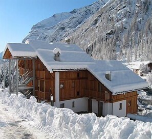 Una cabaña de madera con techo inclinado, rodeada de nieve, con montañas al fondo.