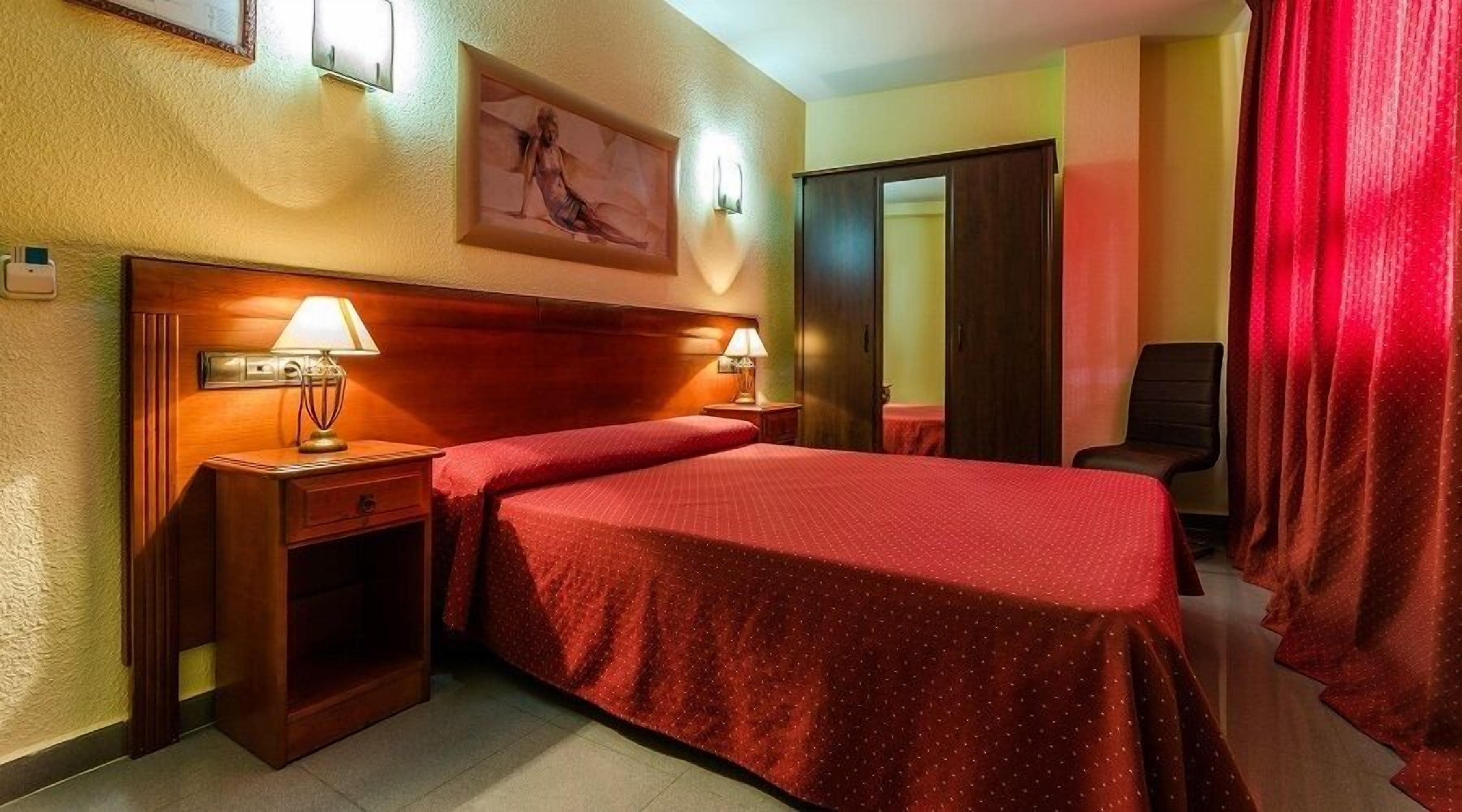 Ein Hotelzimmer mit einem Bett, zwei Nachttischlampen, einem Nachttisch, einem Sessel, einer Tür und einem Bild an der Wand.
