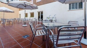 Un patio al aire libre con sillas y mesas de metal, sombrillas y piso de baldosas.