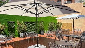 An outdoor patio area with multiple tables and chairs, a large umbrella, and a green wall. - Valdemoro (Valdemoro)