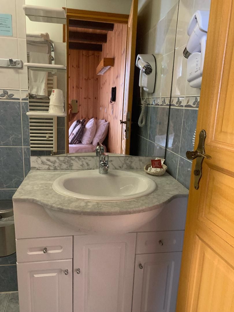 A bathroom with a marble sink, a wooden door, and a mirror reflecting a room with a bed and a wall-mounted hairdryer.