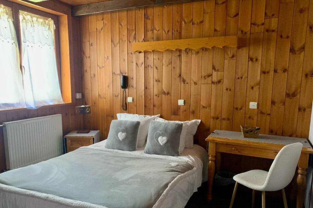 Een slaapkamer met een wand bekleed met houten panelen, een bed met een witte dekbedovertrek, twee kussens met hartmotieven, een houten bureau en een witte stoel.