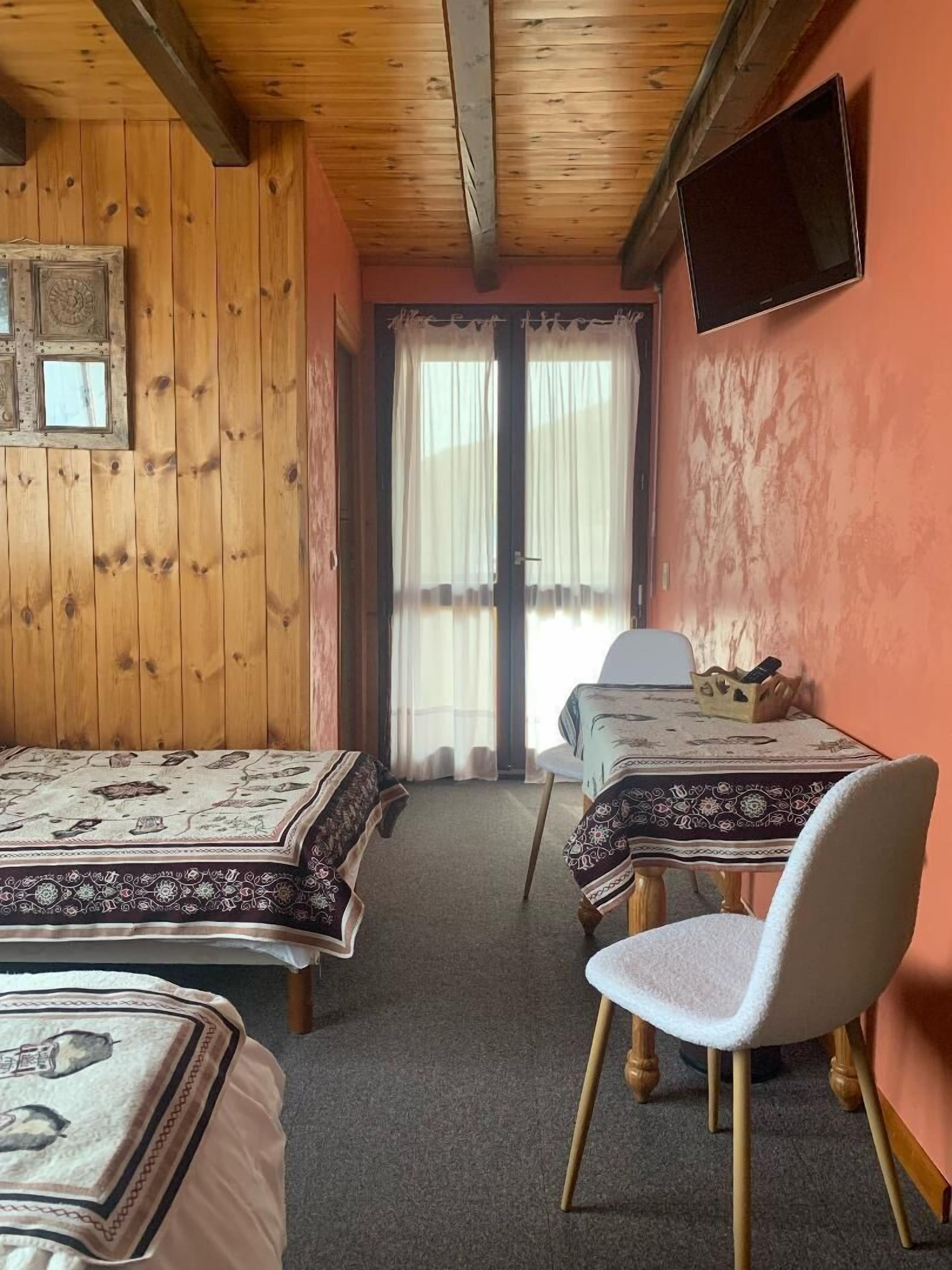 A room with two beds, a table with a tablecloth, a chair, a television mounted on the wall, and a window with curtains.