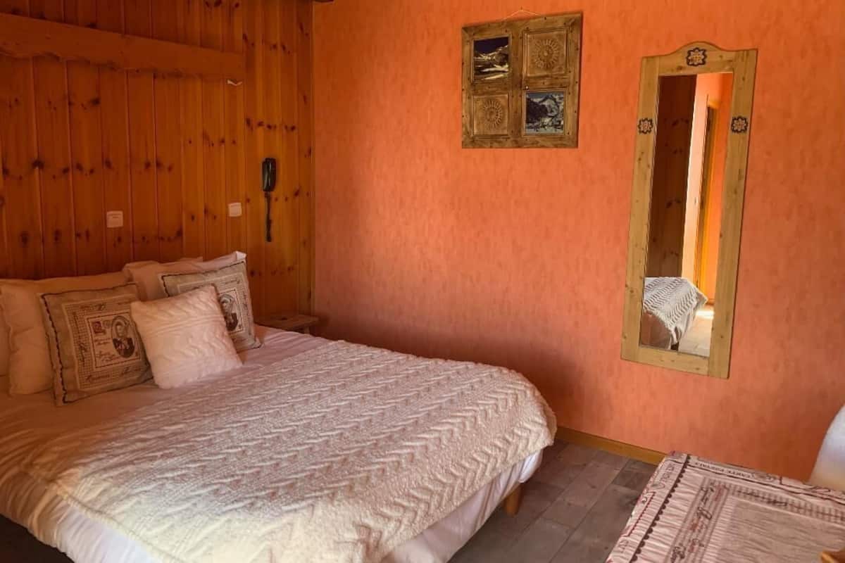 Een slaapkamer met een houten plafond, een bed met behang en een spiegel aan de muur, plus een klein tafeltje met een stoel.