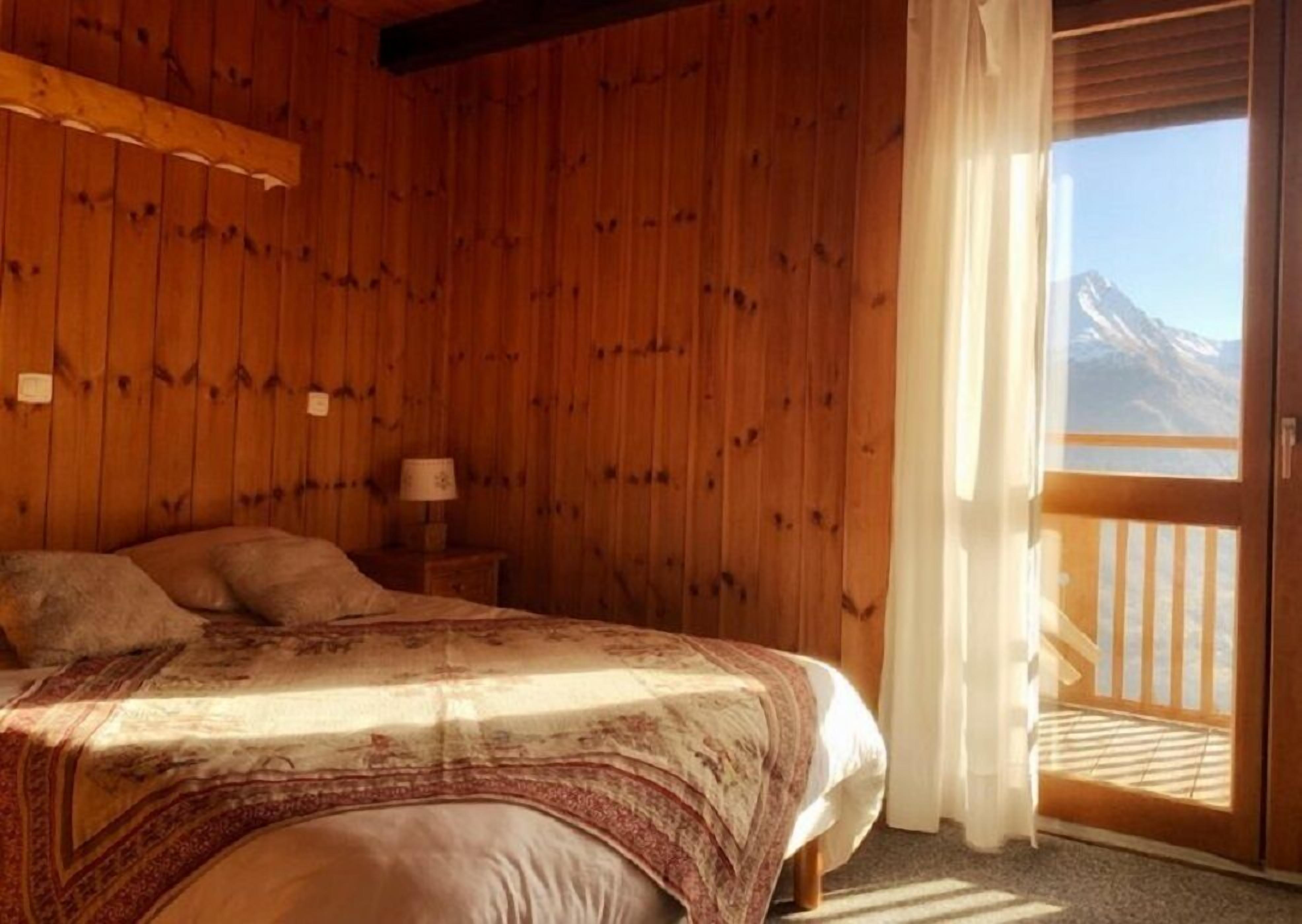 A wooden room with a bed, a nightstand, and a window with a mountain view.
