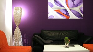 A room with a purple wall, a black leather couch, an orange chair, a white table, and a decorative glass vase. - Apartment Berlin 1 (Berlin)