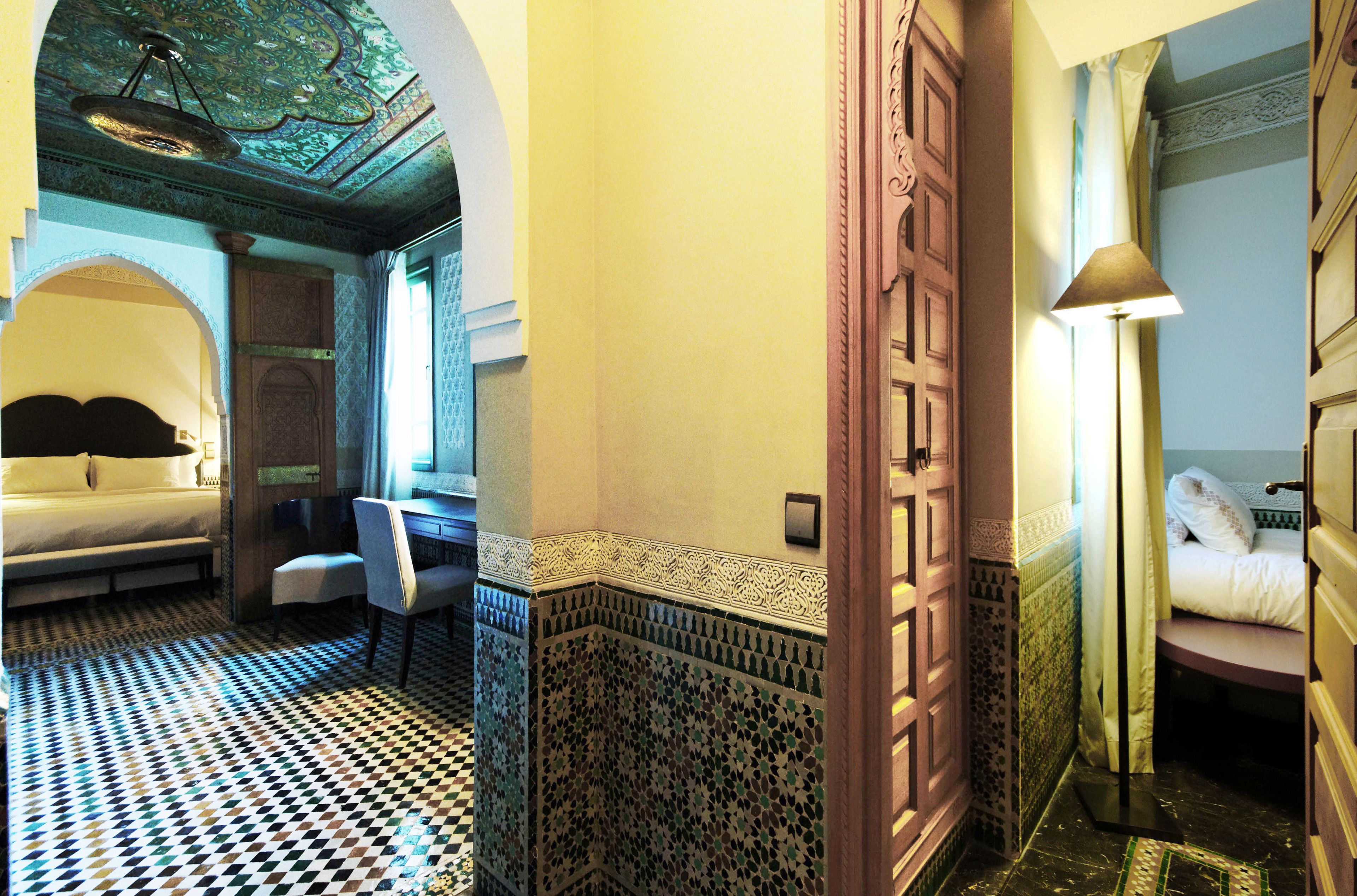 A hotel room with a checkered floor, a bed, a desk, and a bathroom with patterned tiles.