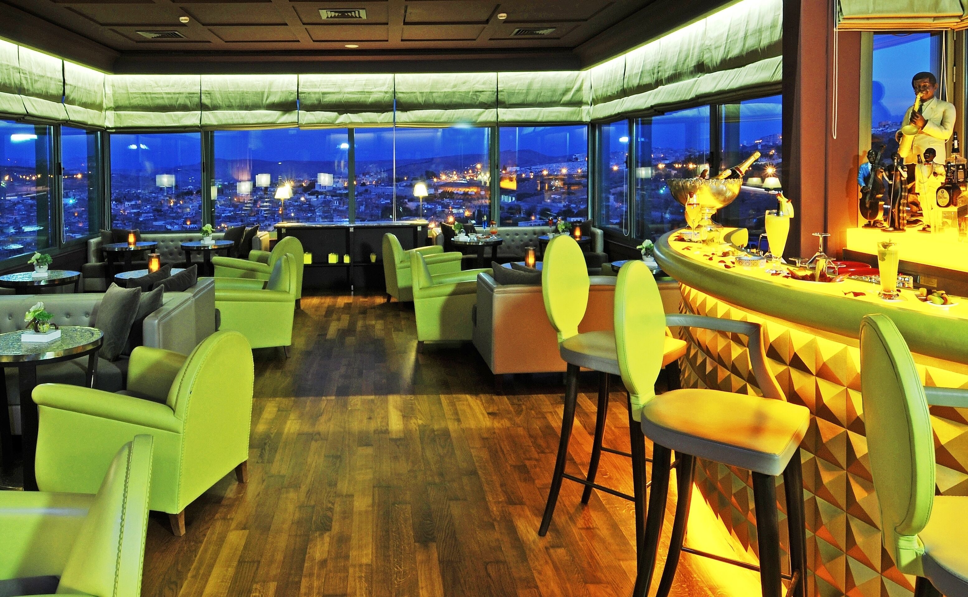 A restaurant with a city view, green chairs, and a bar area.