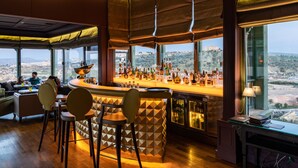 Een bar met uitzicht op een stadsgezicht door grote ramen, een toonbank met flessen en barstoelen.