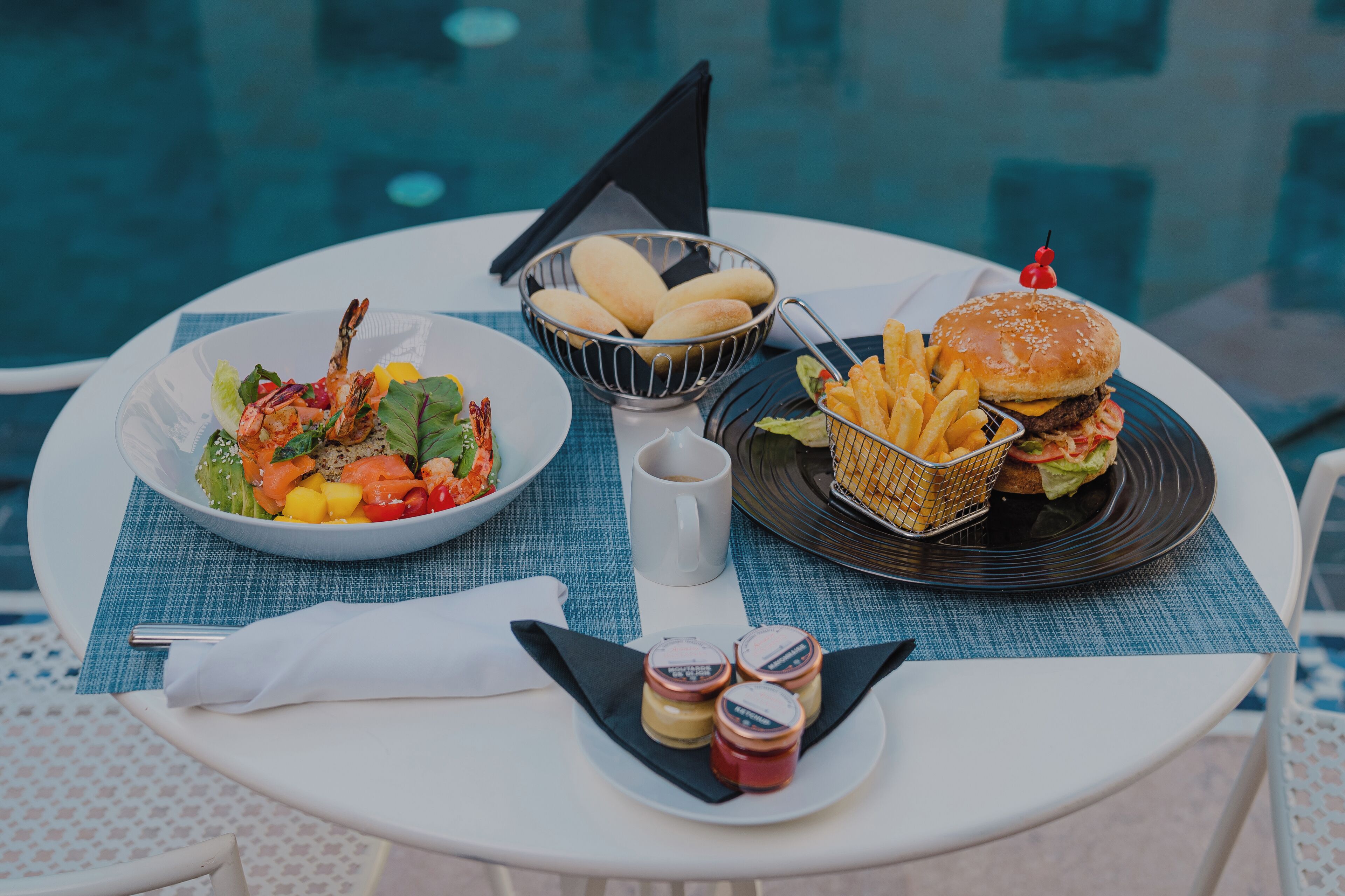 Una mesa de comedor con ensalada, pan, una hamburguesa, papas fritas y salsas al lado de una piscina.
