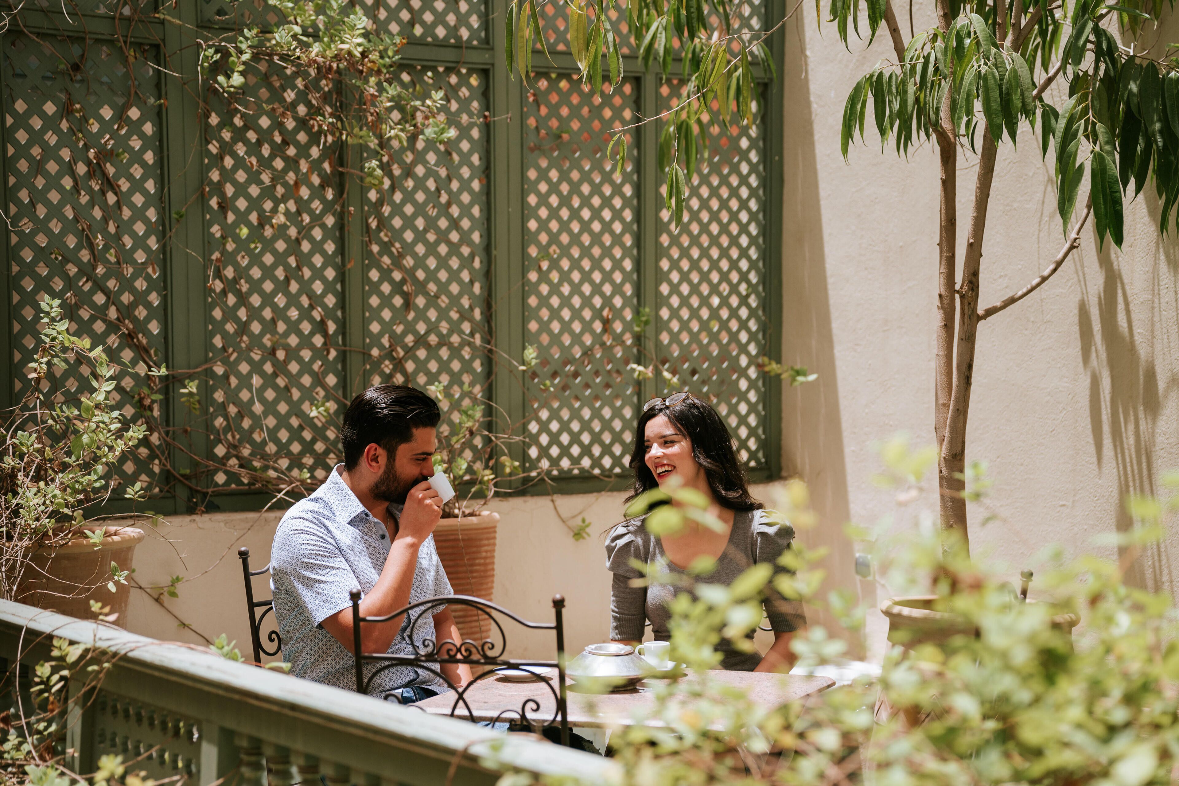 Un couple est assis à une table à l’extérieur, avec une clôture en treillis et des plantes en arrière-plan.