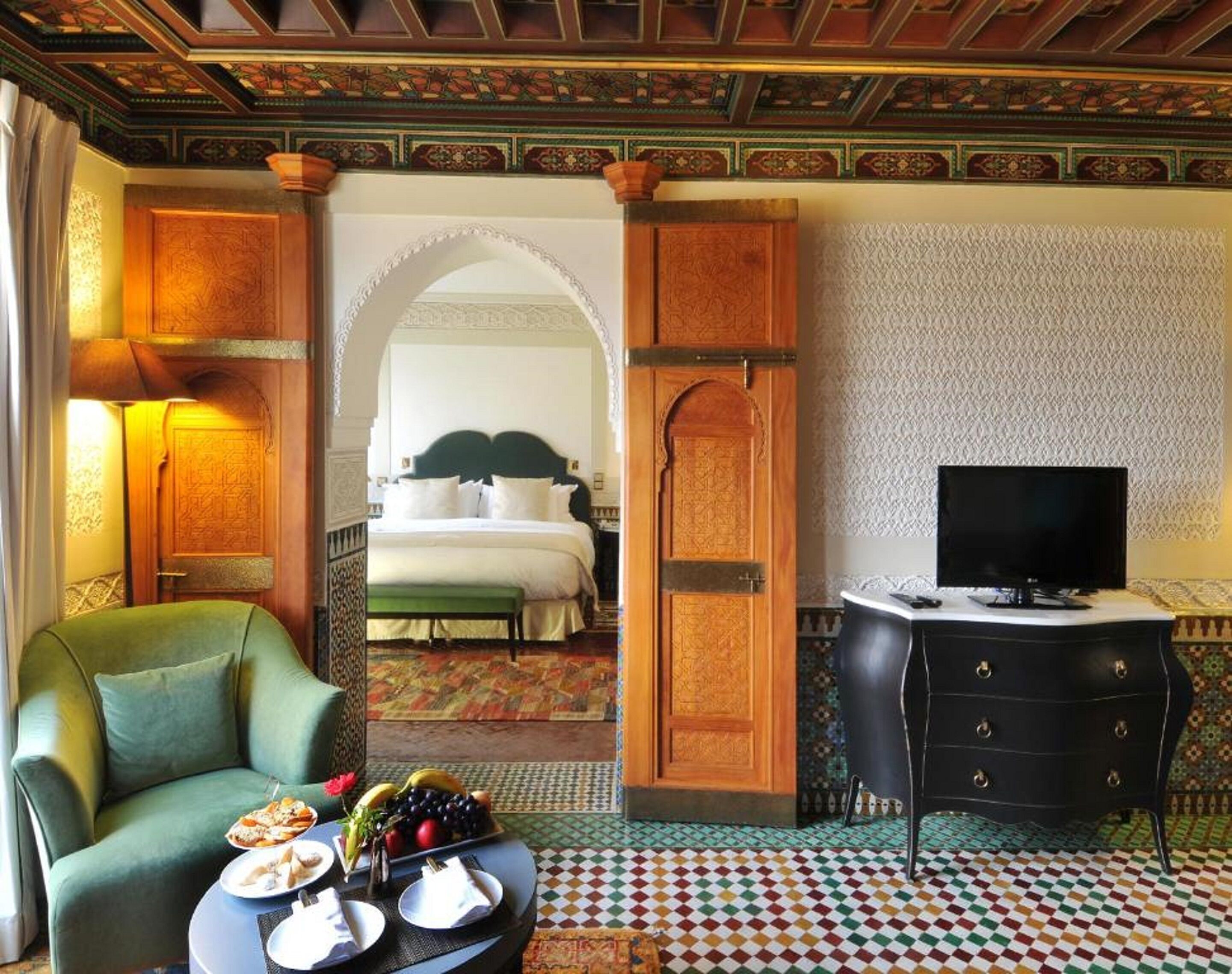 Une chambre d’hôtel avec un lit, une télévision, un fauteuil vert et une table à manger avec des assiettes et un panier de fruits.