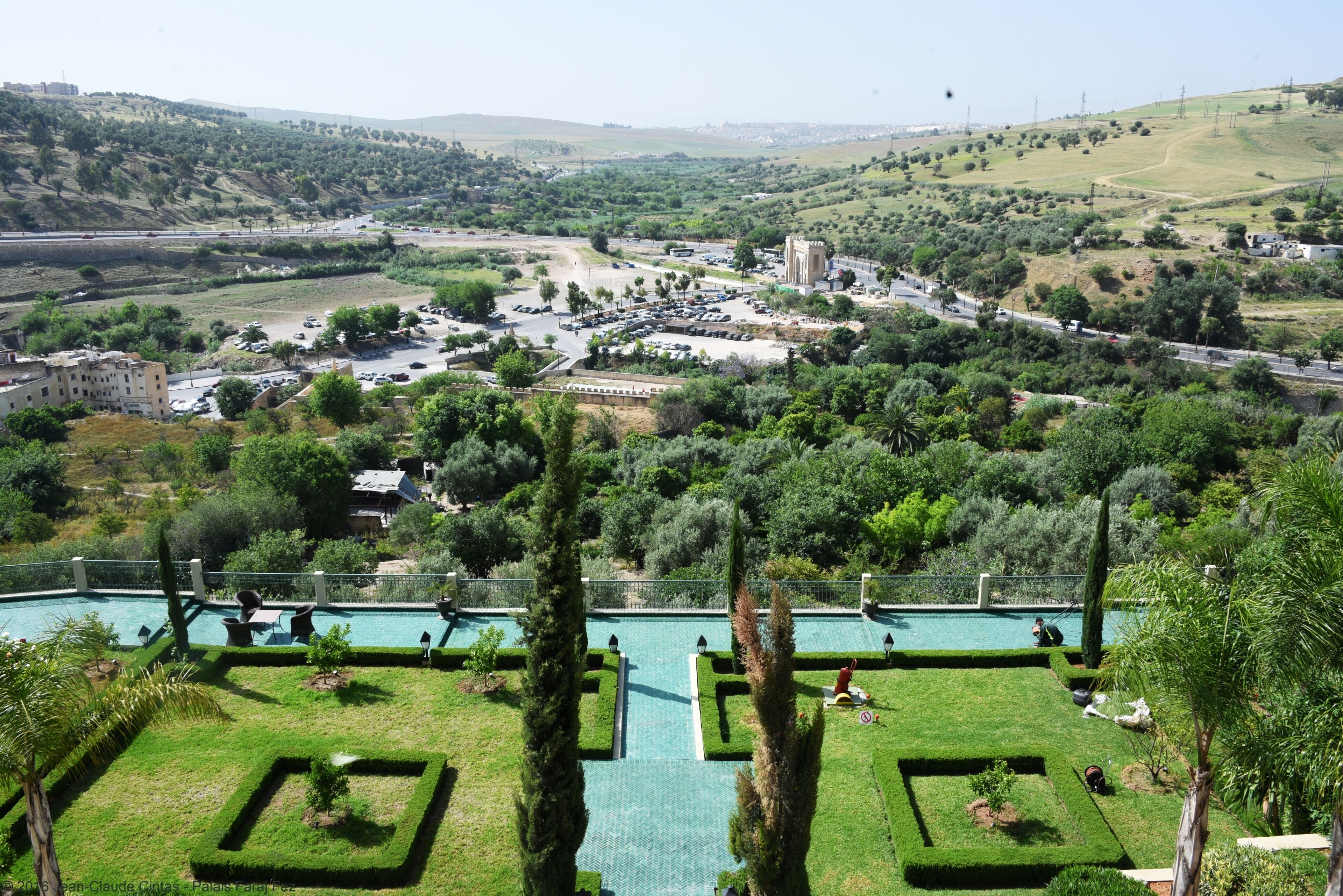 A pool area with geometric landscaping, a view of a town with buildings and roads, and a hilly landscape in the distance.