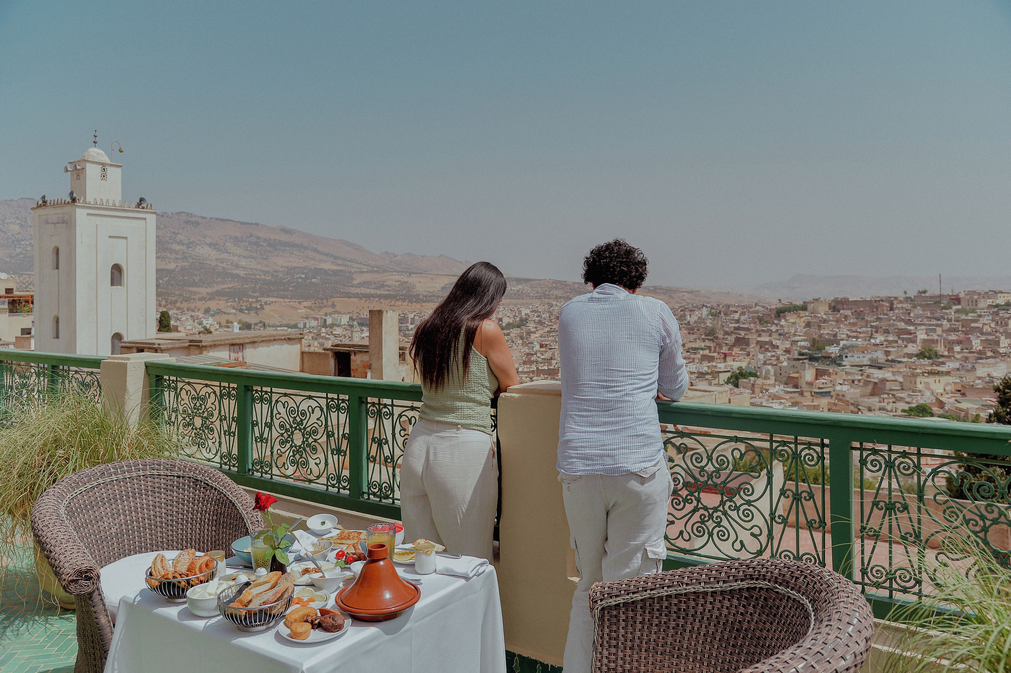 Varanda com cadeiras de vime, uma mesa com comida e vista para a paisagem urbana e uma torre branca.