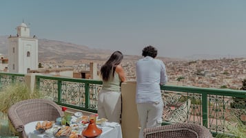 Varanda com cadeiras de vime, uma mesa com comida e vista para a paisagem urbana e uma torre branca.