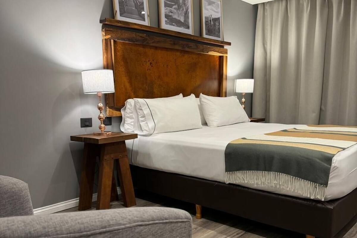 A modern hotel room with a large bed, wooden headboard, and a grey wall. There are two bedside tables with lamps, a grey armchair, and a wooden floor.