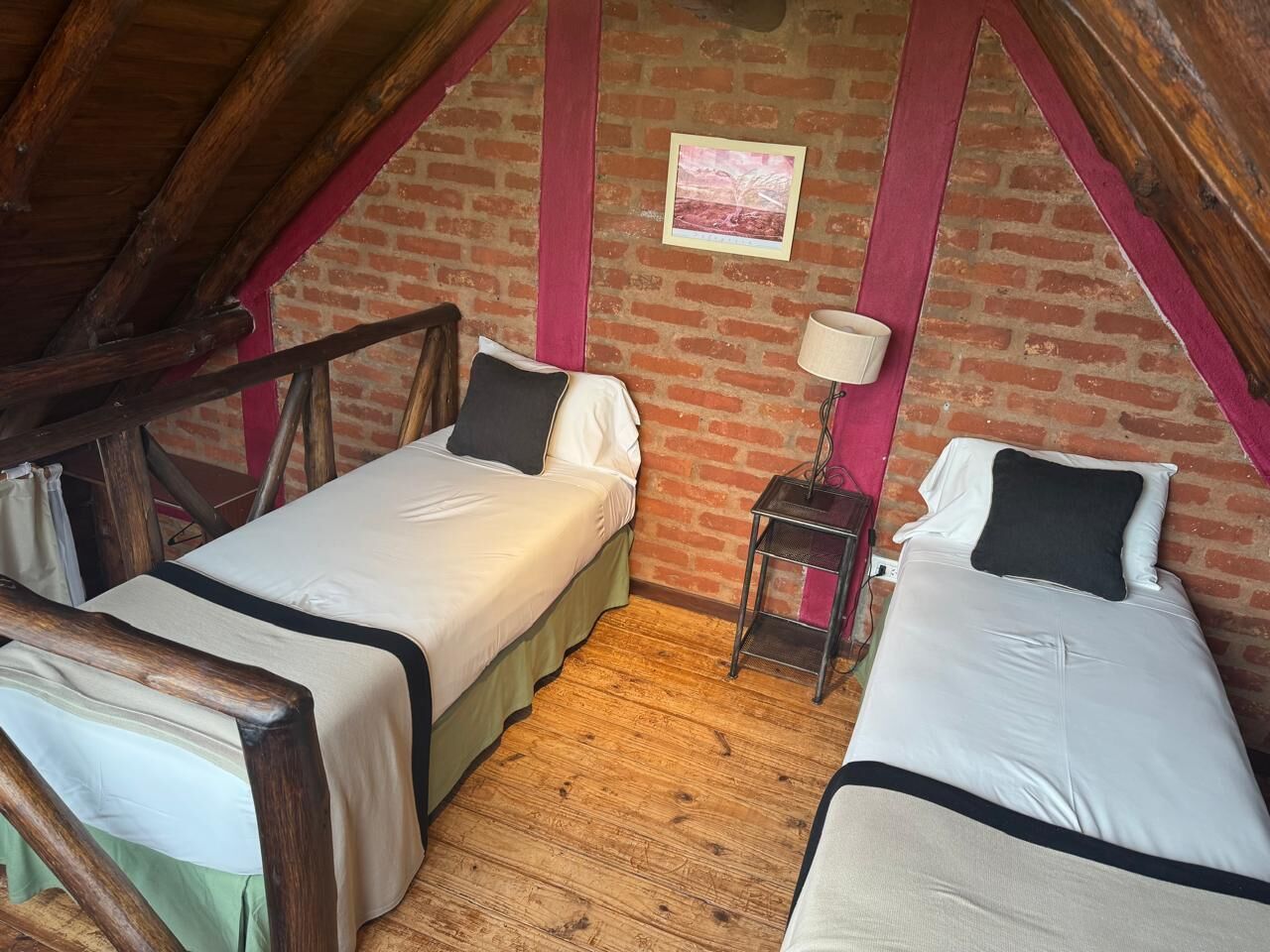 Ein kleiner, rustikaler Raum mit zwei Einzelbetten, einem Holzstuhl, einem Tisch und einer Ziegelwand mit einem Bild.