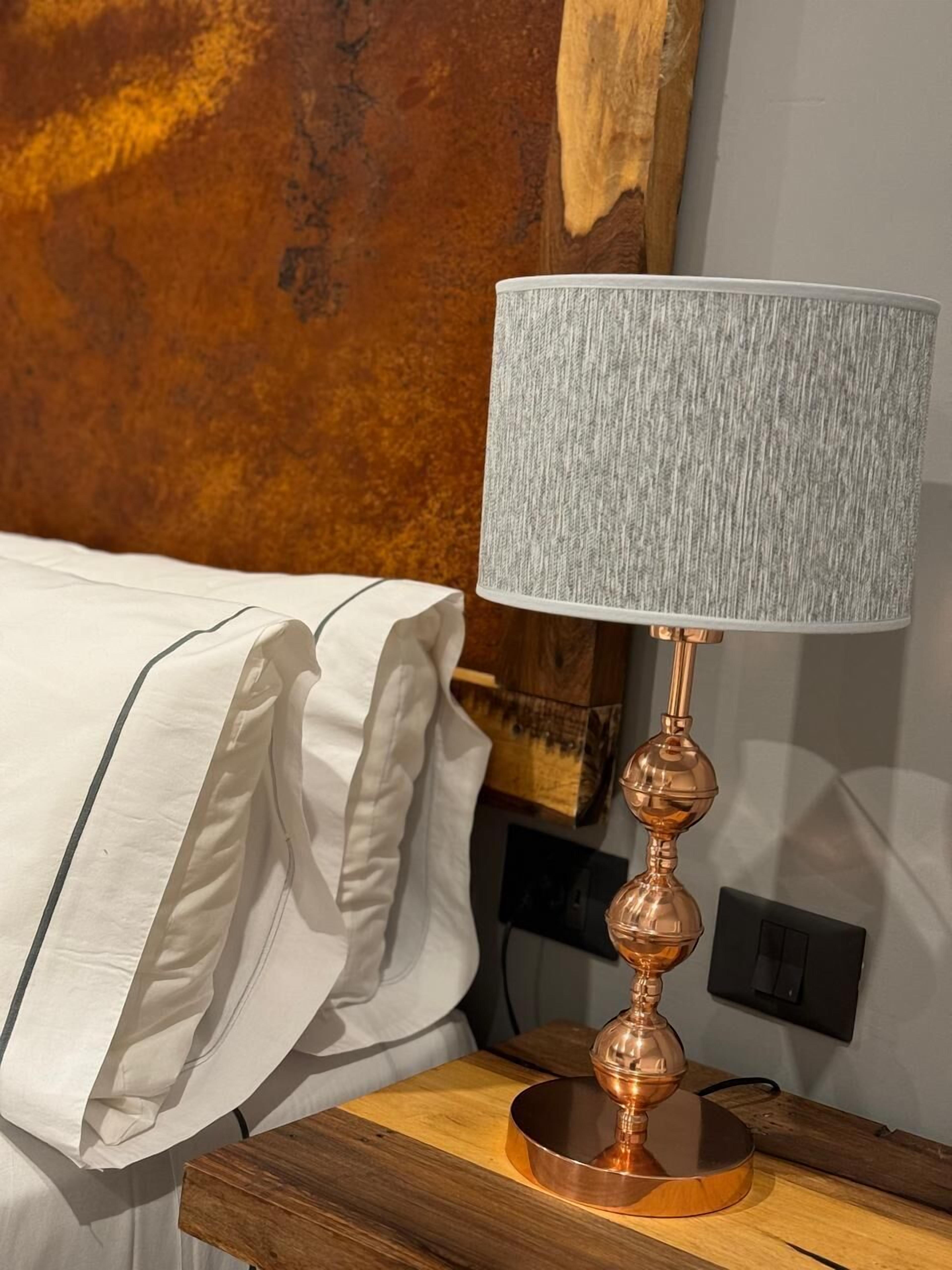 Un lit bien fait, avec du linge de lit blanc et une lampe grise à texture sur une table de chevet en bois.