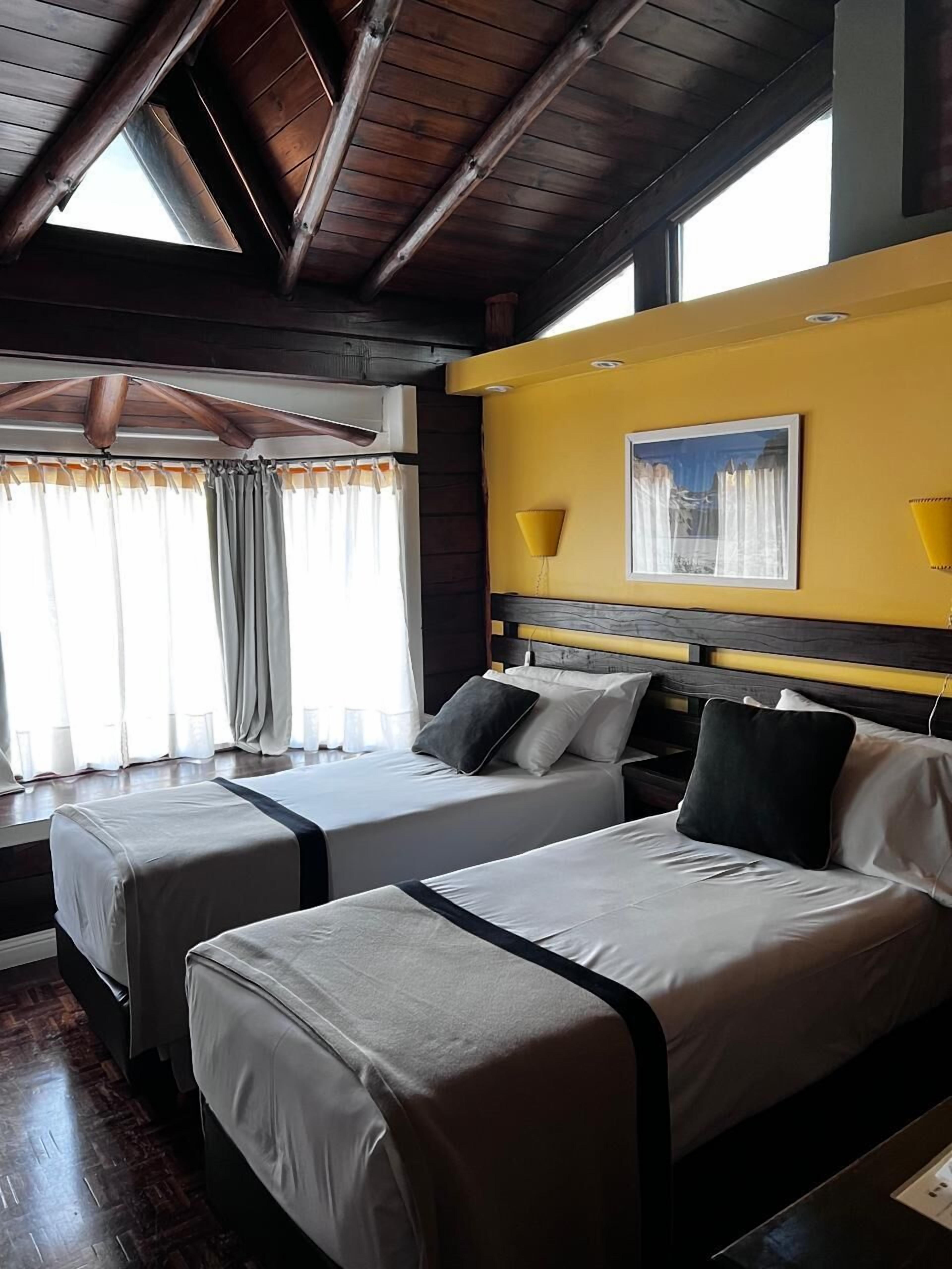 A room with two beds, wooden beams on the ceiling, a yellow wall, and a window with curtains.