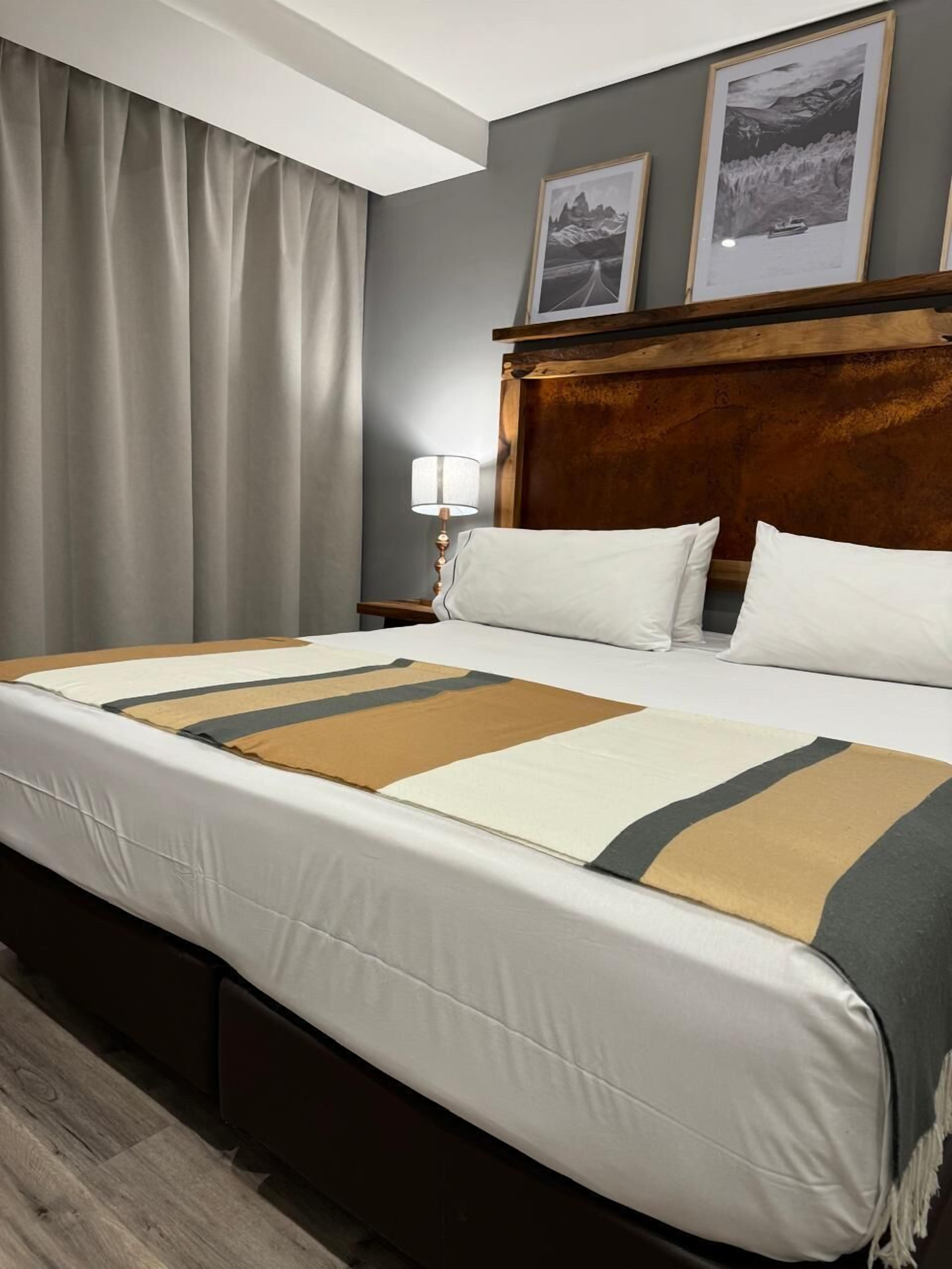 Ett hotellrum med en stor säng, grå gardiner, en sänggavel i trä och två inramade tavlor på väggen.