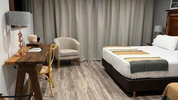A hotel room with a wooden floor, a bed with a white and brown bedspread, a desk with a lamp, a chair, and a large window with curtains.