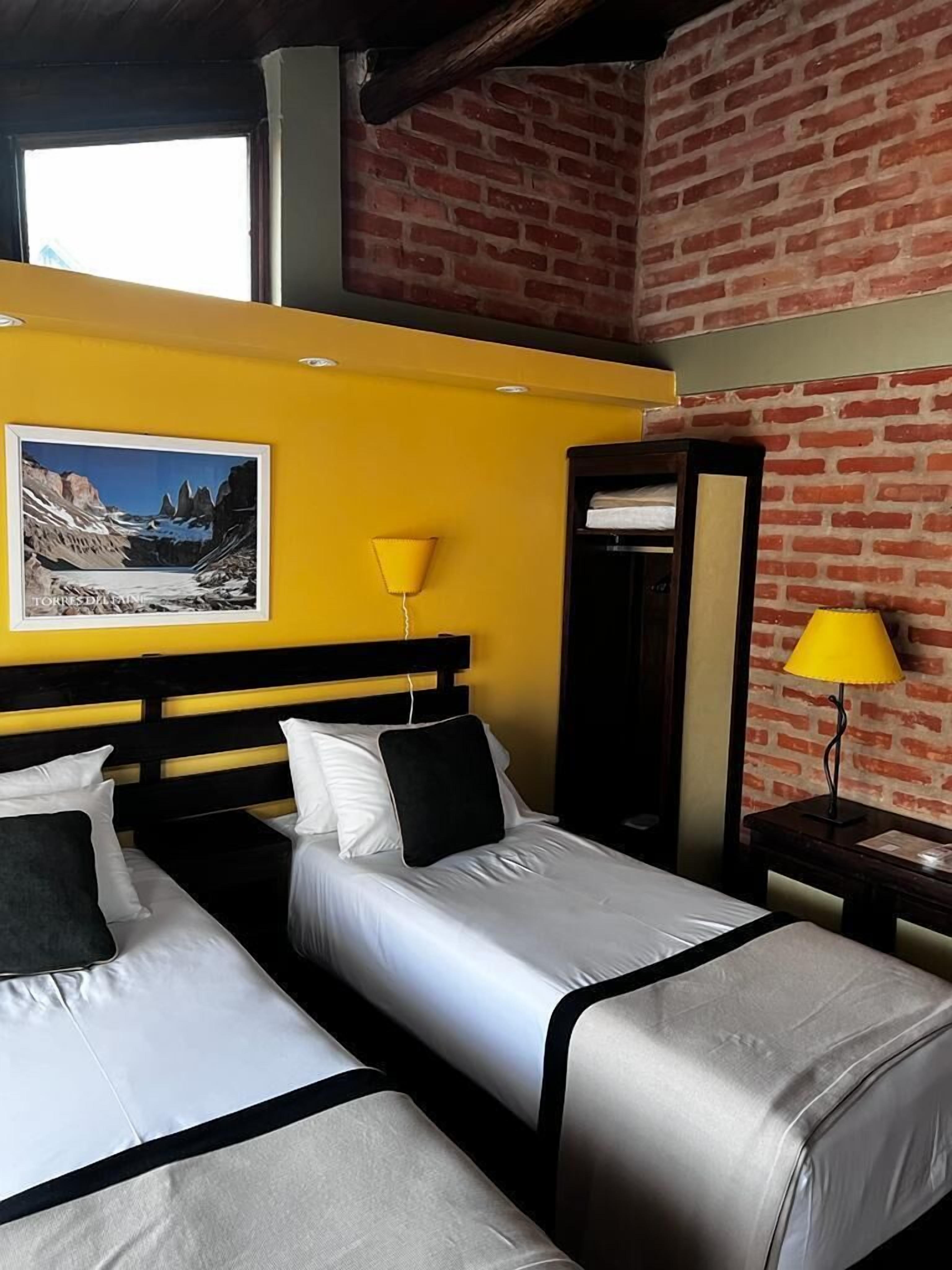 Habitación de hotel con dos camas, una pared amarilla, una pared de ladrillo y un cuadro enmarcado en la pared.