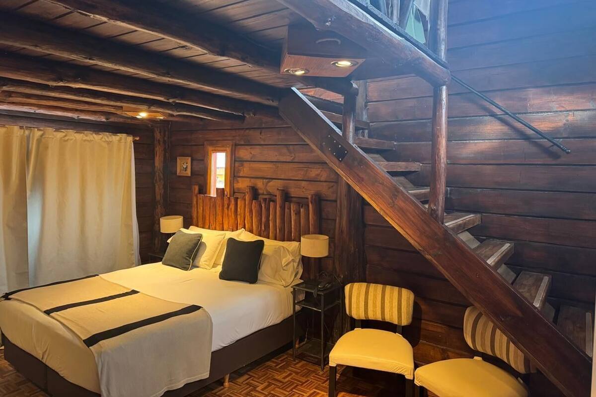 A wooden cabin bedroom with a bed, two chairs, a nightstand, and a staircase.