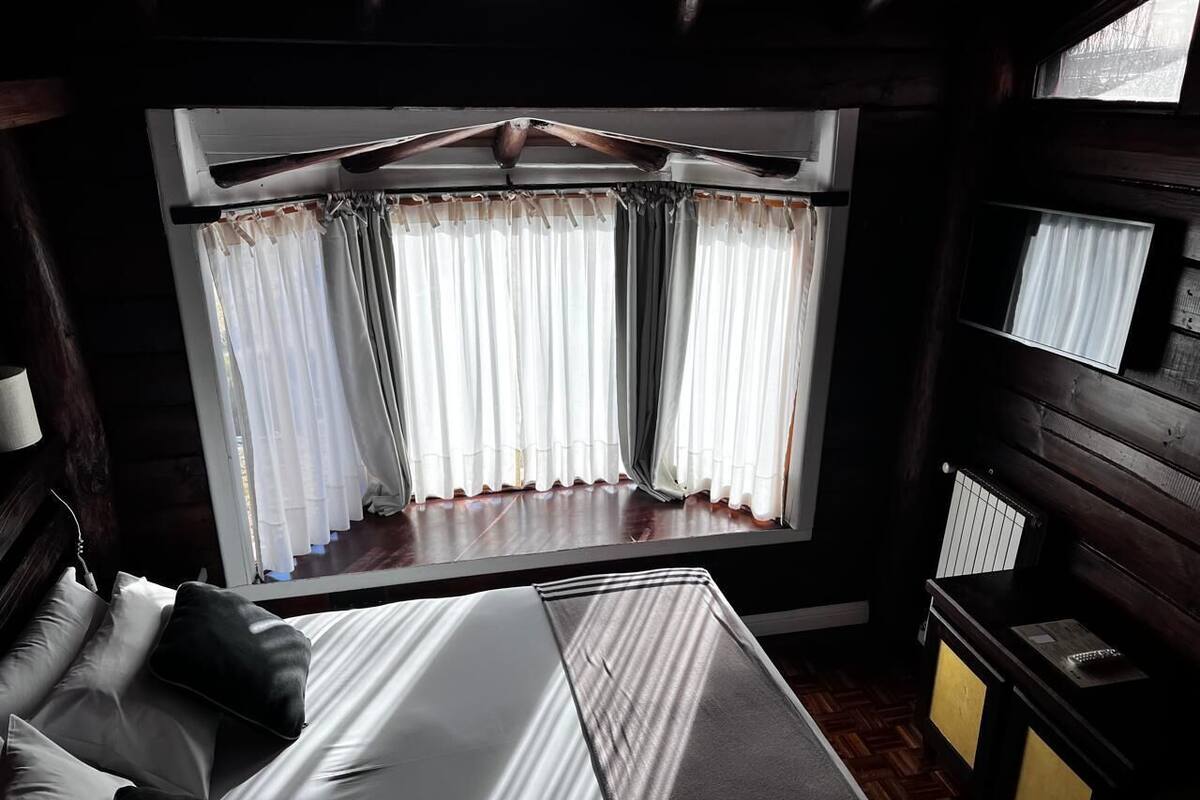 A bedroom with a large window, a bed with white and gray linens, a wooden headboard, and a bedside table with a lamp.
