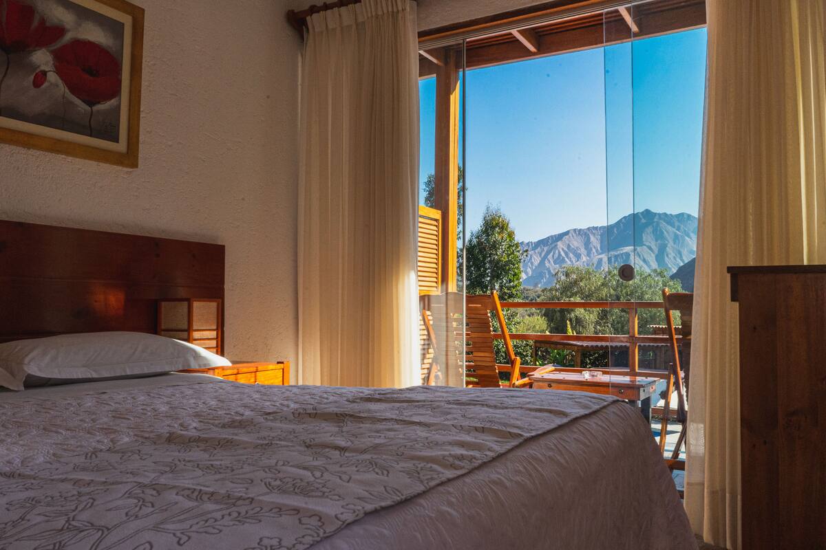Ein Schlafzimmer mit einem großen Bett, einem hölzernen Kopfteil, einem Gemälde und Bergblick durch eine Glastür.
