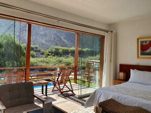 A hotel room with a large window offering a mountain view, a bed, a wooden table, and a chair.