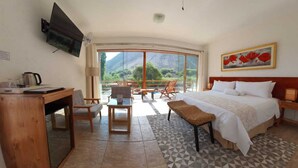 Une chambre d’hôtel équipée d’un lit, d’un bureau, d’une chaise, d’une table et d’un téléviseur. Une grande fenêtre offre une vue sur les montagnes.