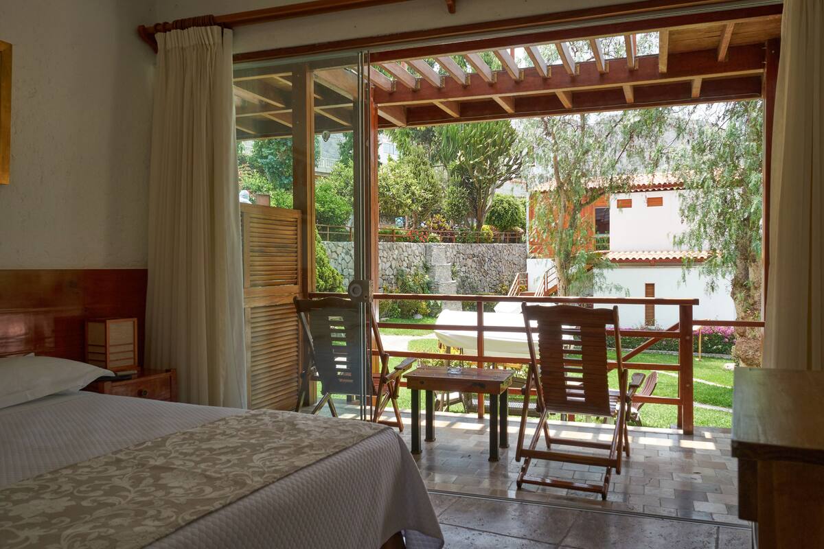 Ein Hotelzimmer mit Bett, Holzstuhl, kleinem Tisch und Blick auf Garten und Gebäude.