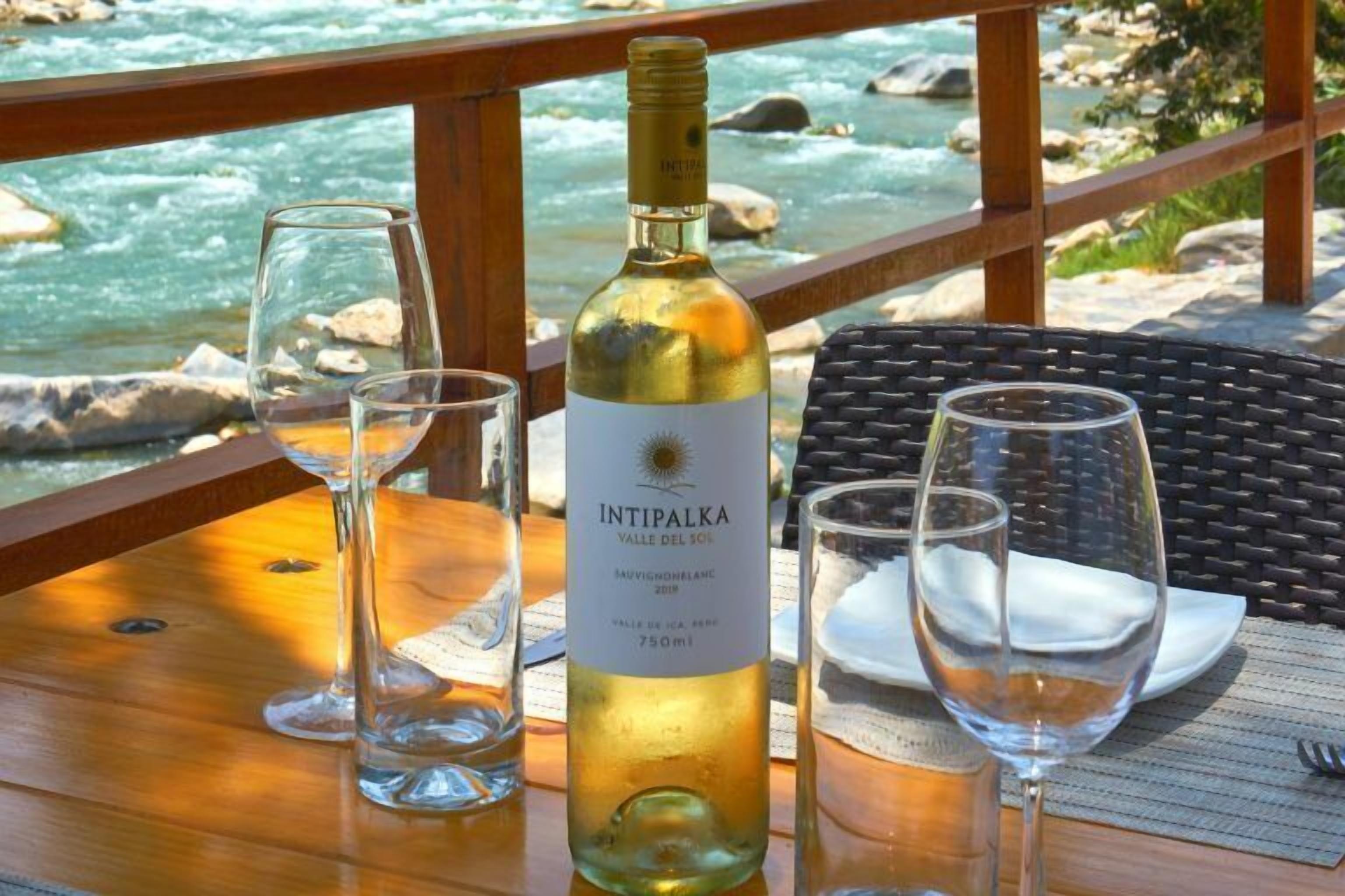 Una botella de vino Intipalka sobre una mesa de madera, con copas y un plato, con vista a un río.