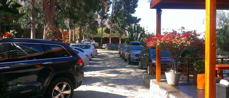 A parking area with several parked cars, a covered seating area with potted plants, and a pathway lined with trees.
