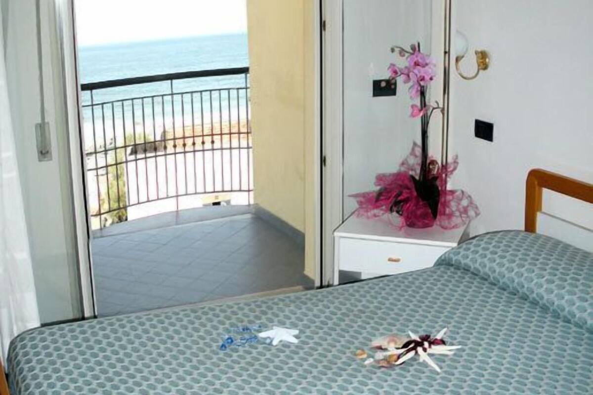 Une chambre à coucher avec un lit, une table de chevet, un vase de fleurs et une vue sur la mer, visible par la porte d’un balcon.