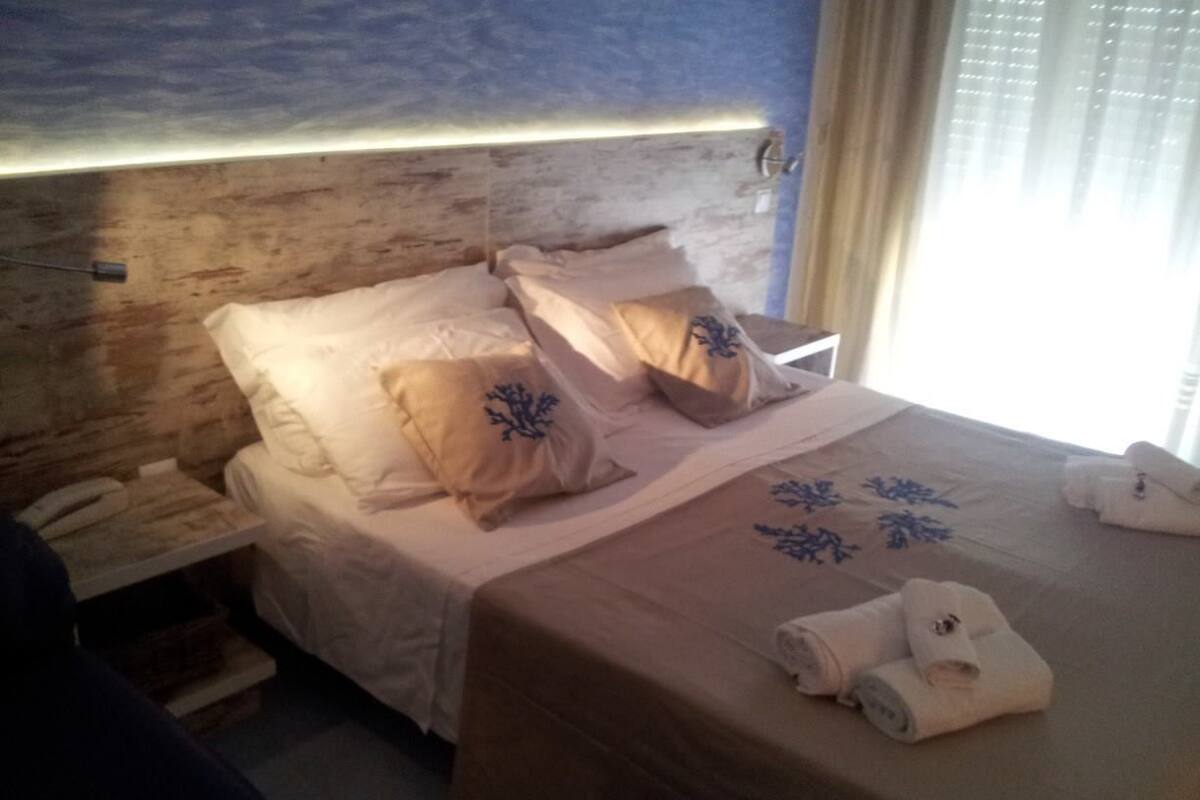 Une chambre d’hôtel avec une tête de lit en bois, un lit avec une literie beige et des coussins à motifs floraux bleus, une table de chevet avec une lampe et une fenêtre avec des rideaux.