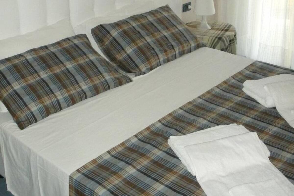 Une chambre d’hôtel avec un grand lit, deux oreillers, une table de chevet avec une lampe et une fenêtre avec des rideaux légers.