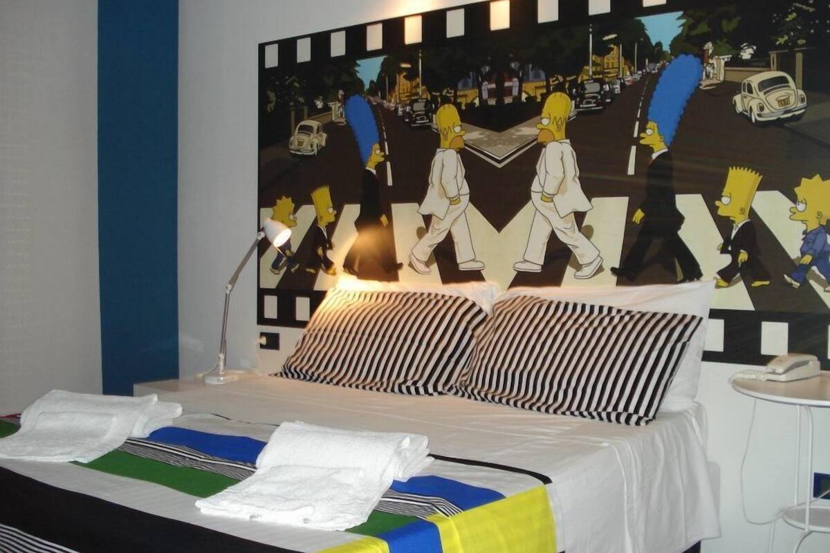 Une chambre d’hôtel avec un lit, une table de chevet et une grande œuvre d’art sur le mur, dont le thème est celui des Simpsons.