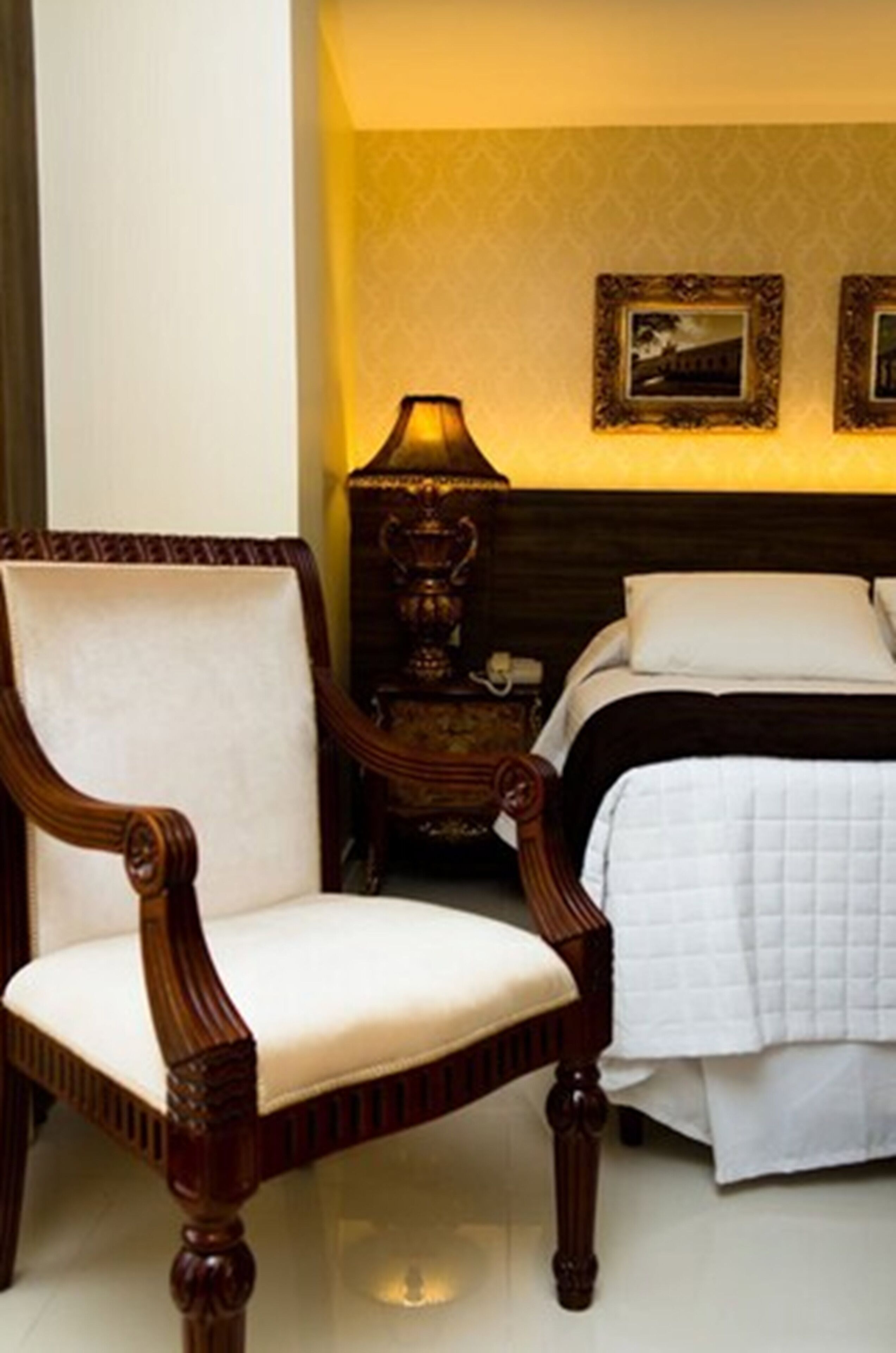 A bedroom with a wooden chair, a bedside table with a lamp, and a bed with white bedding.