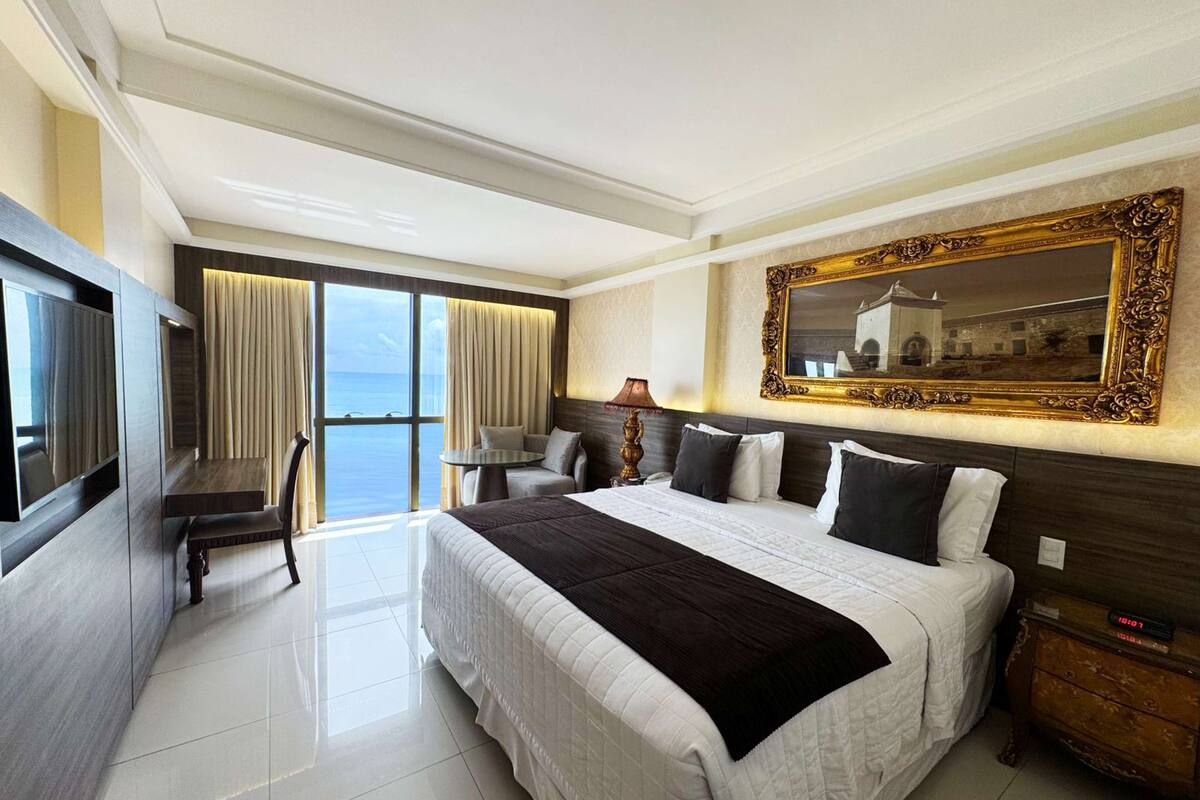 A hotel room with a large bed, a TV, a desk, and a view of the sea.